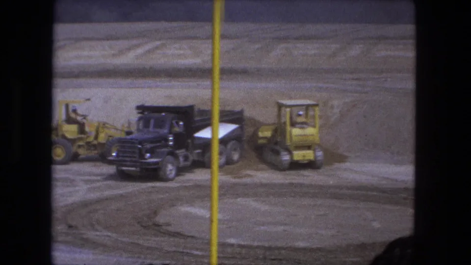 A black dump truck is flanked by yellow construction equipment on a dusty 1974 Fort Wayne worksite.
