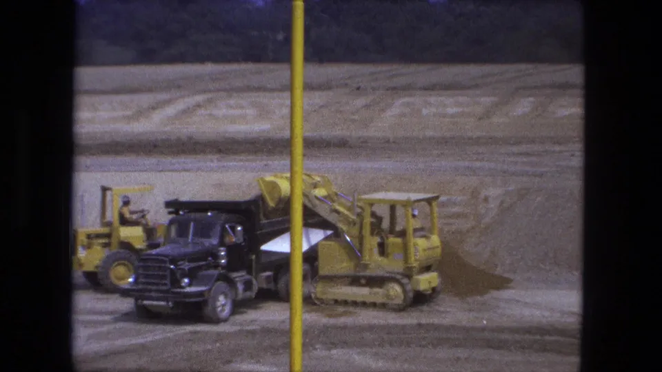 1974 Fort Wayne 8mm frame showing a yellow loader near a black dump truck at an open construction excavation.