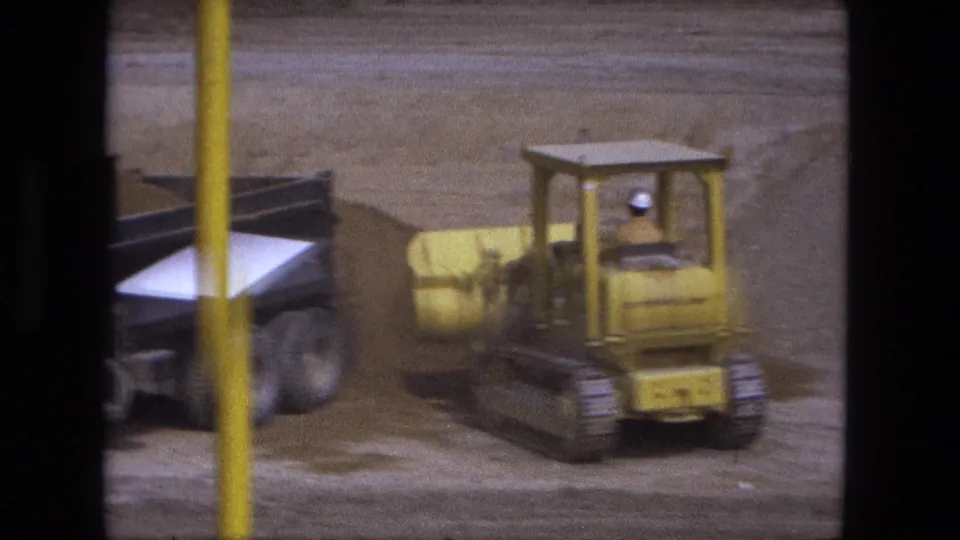 A yellow loader passes close to a dump truck at the Fort Wayne excavation site, with film grain and a foreground pole visible.