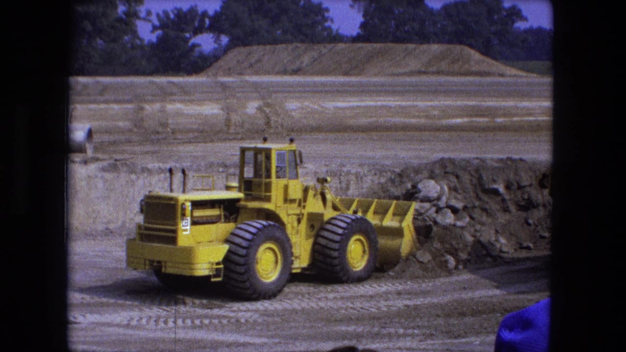 Yellow front loader approaches a pile of dirt and rock on a graded lot.