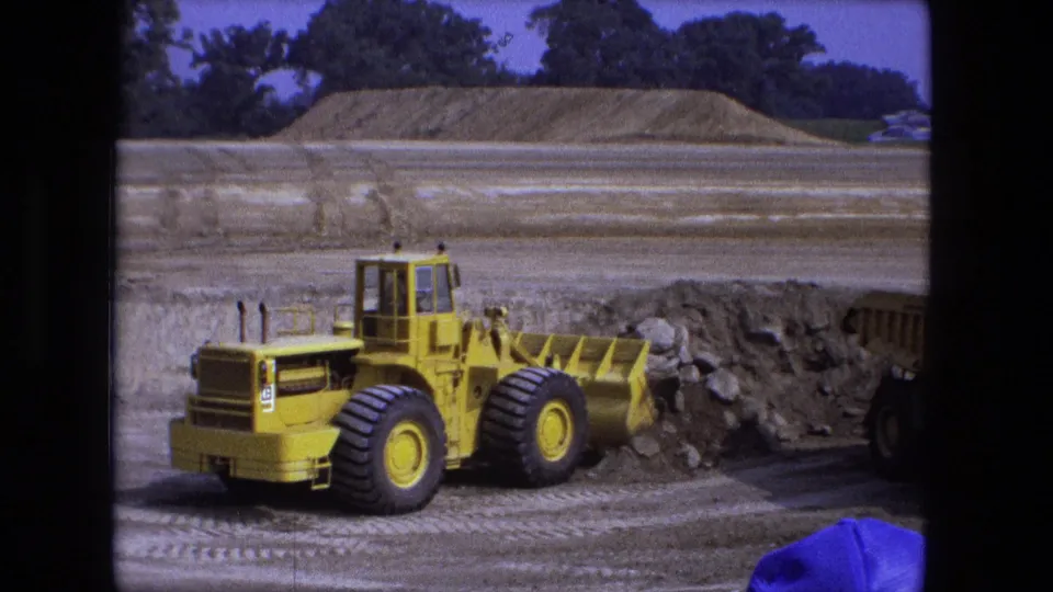 1974 construction-site frame with a yellow loader, soil mound, tire tracks, and a dump truck partly entering from the right.