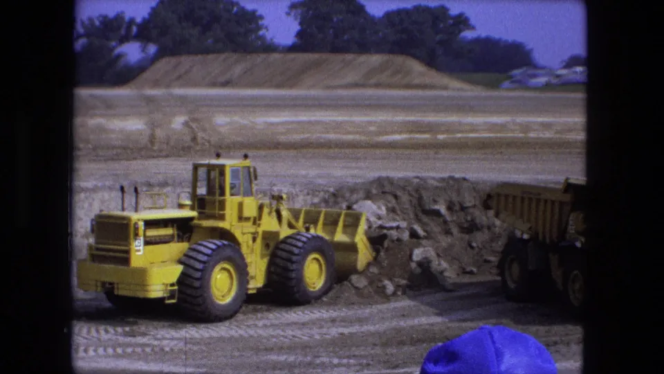 Fort Wayne construction frame showing the yellow loader beside excavated soil, with a dump truck partly visible on the right edge.