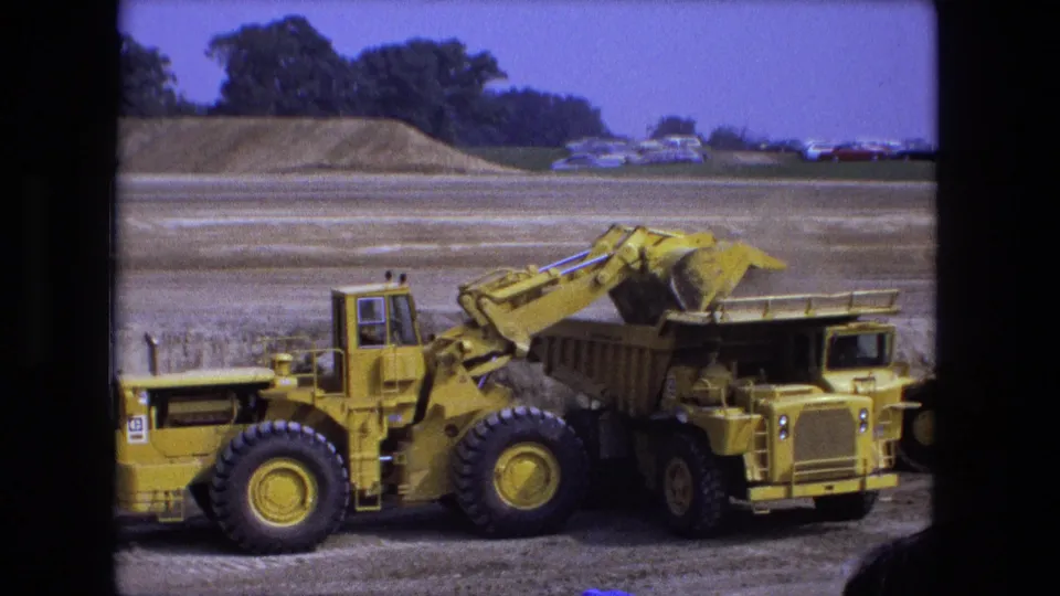 A yellow front-end loader continues loading a dump truck in Fort Wayne, with earth piles and parked cars in the background.