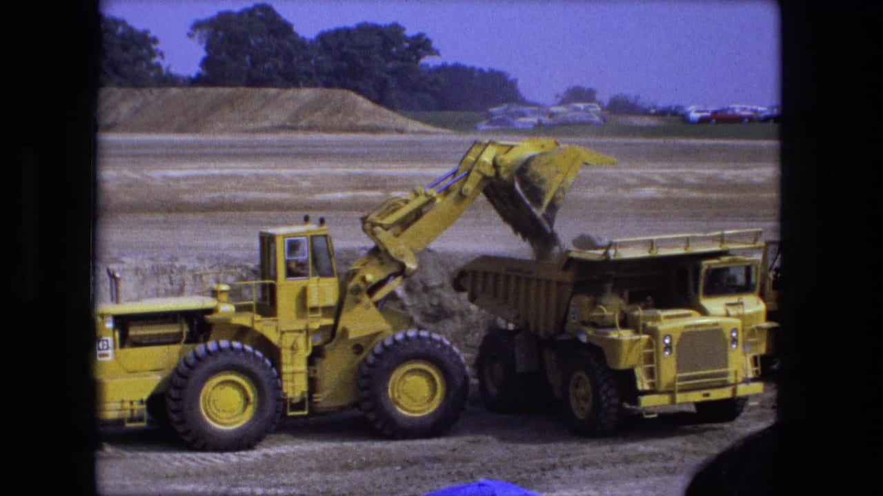 Yellow front-end loader dumps dirt into a yellow dump truck at an open worksite.