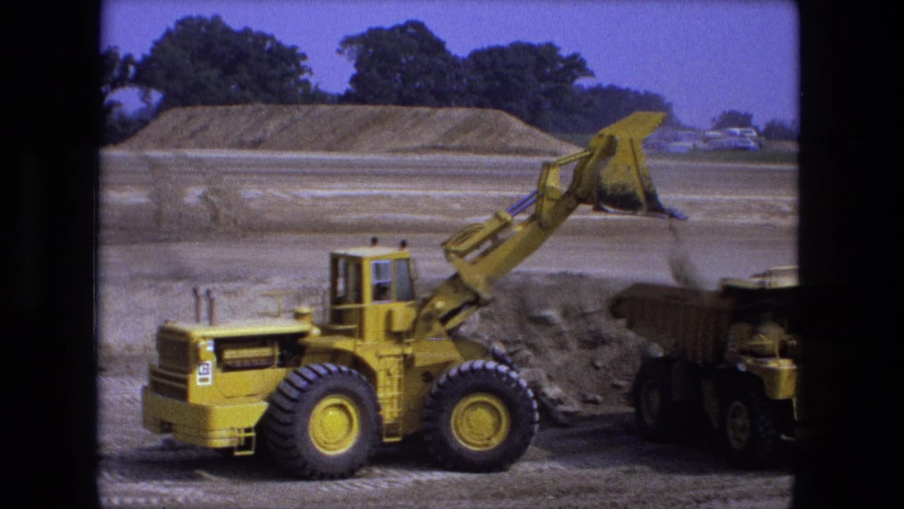 1974 Fort Wayne frame with a yellow loader finishing a dump into a truck on a wide, dusty construction site.