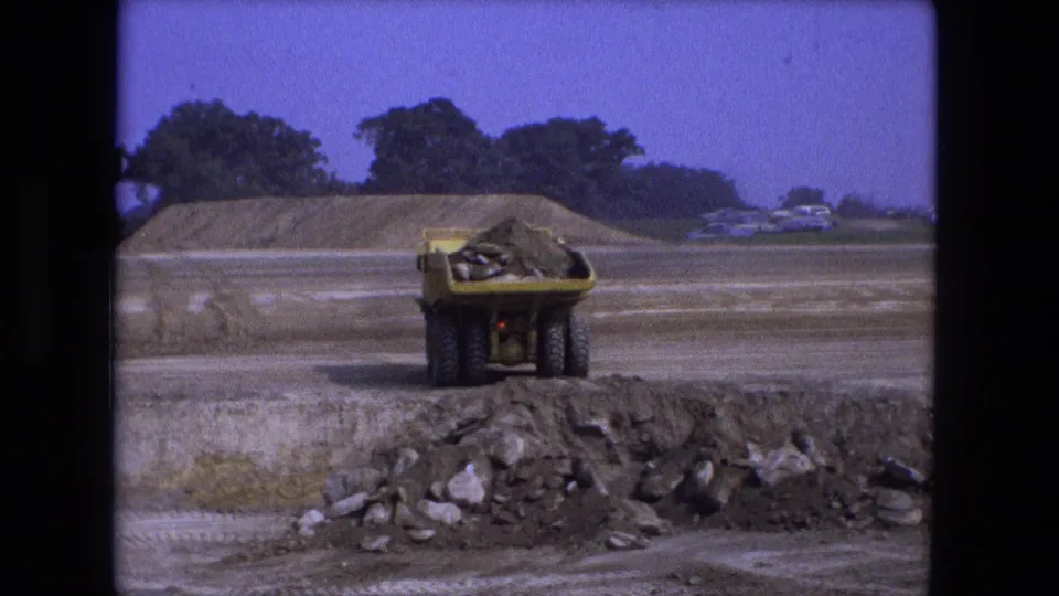 1974 Fort Wayne frame of a loaded yellow dump truck receding across the excavation area under a blue sky.