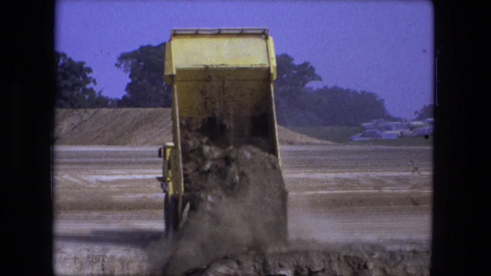 A yellow dump truck finishes emptying dirt and rocks onto a dusty Fort Wayne construction surface in 1974.