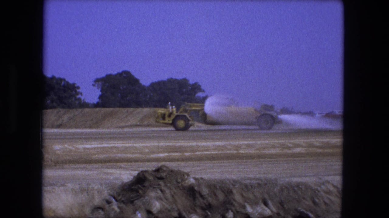 1974 Fort Wayne 8mm frame showing a yellow water truck spraying water across a dirt construction road under a purple-blue sky.
