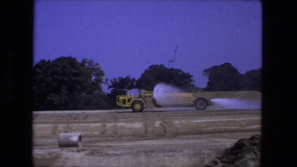 A 1974 home-movie frame shows the water truck farther across the dirt road, with trees behind and a small cylinder-like object in front.