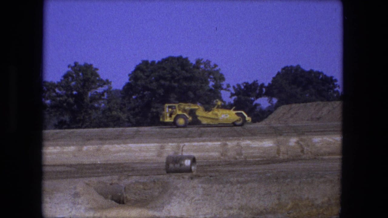 Yellow road grader moves across a dirt construction site with trees behind it.
