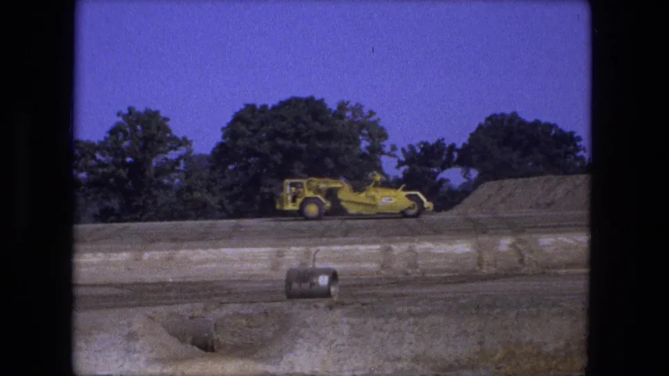 A yellow road grader crosses a Fort Wayne, Indiana dirt construction site in 1974, with earth piles and trees in the background.