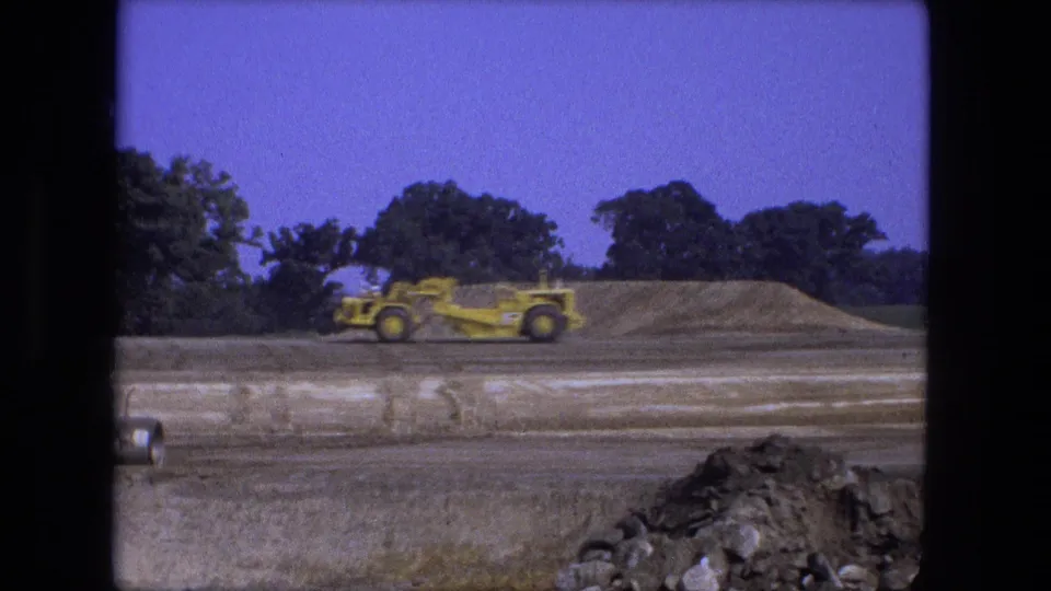 The grader moves past a long soil pile at the Fort Wayne construction site, framed by trees and a blue 8mm sky.