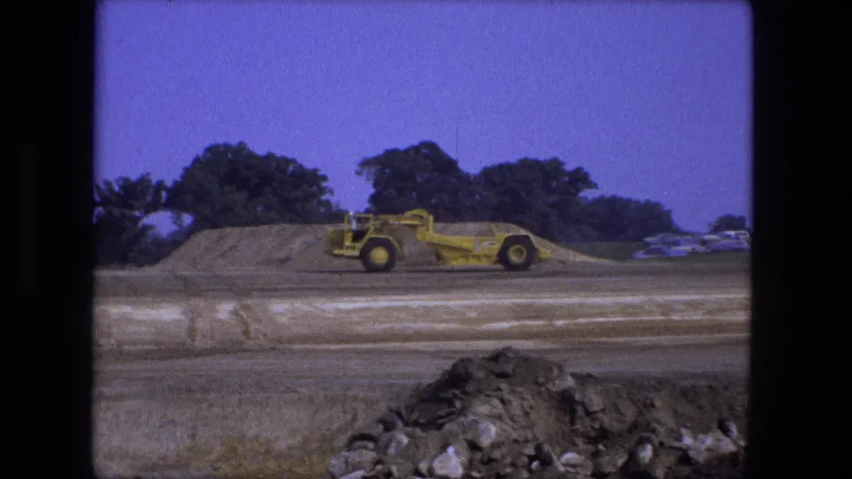 A yellow road grader is seen broadside at the 1974 Fort Wayne site, with a mound, foreground rocks, trees, and distant cars visible.