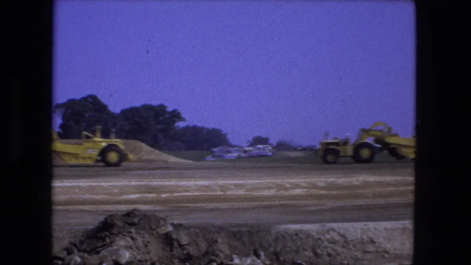 1974 Fort Wayne 8mm frame showing a yellow road grader moving across a graded dirt site with trees and parked cars in the background.