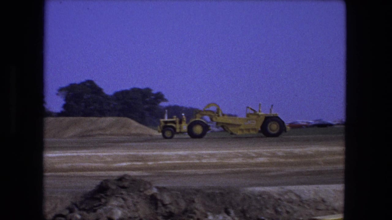 Yellow road grader in profile on a dirt site with trees, cars, and a low mound behind it.