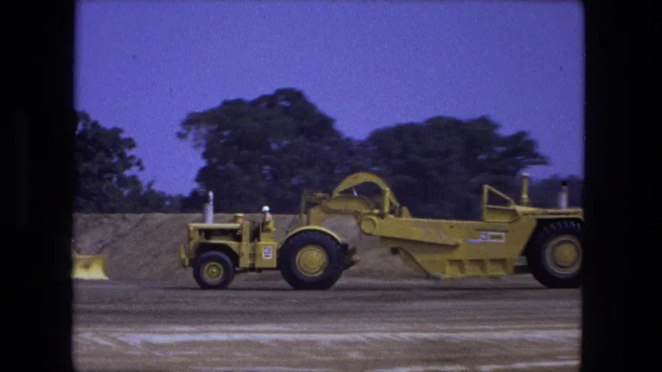 Closer 1974 Fort Wayne 8mm view of the yellow grader, with its cab area and operator position visible against trees and dirt.
