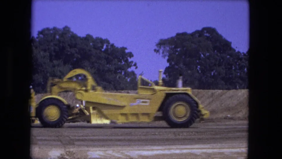 1974 Fort Wayne frame showing the yellow road grader close to the camera, with blade, cab structure, trees, and graded earth visible.