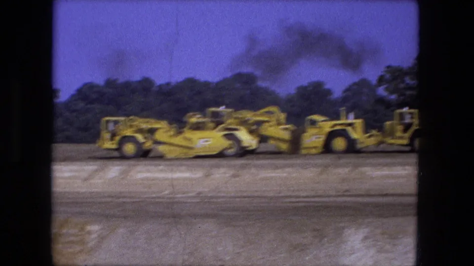 1974 Fort Wayne 8mm frame showing yellow earth-moving equipment across a scraped dirt site with trees and dark smoke in the background.
