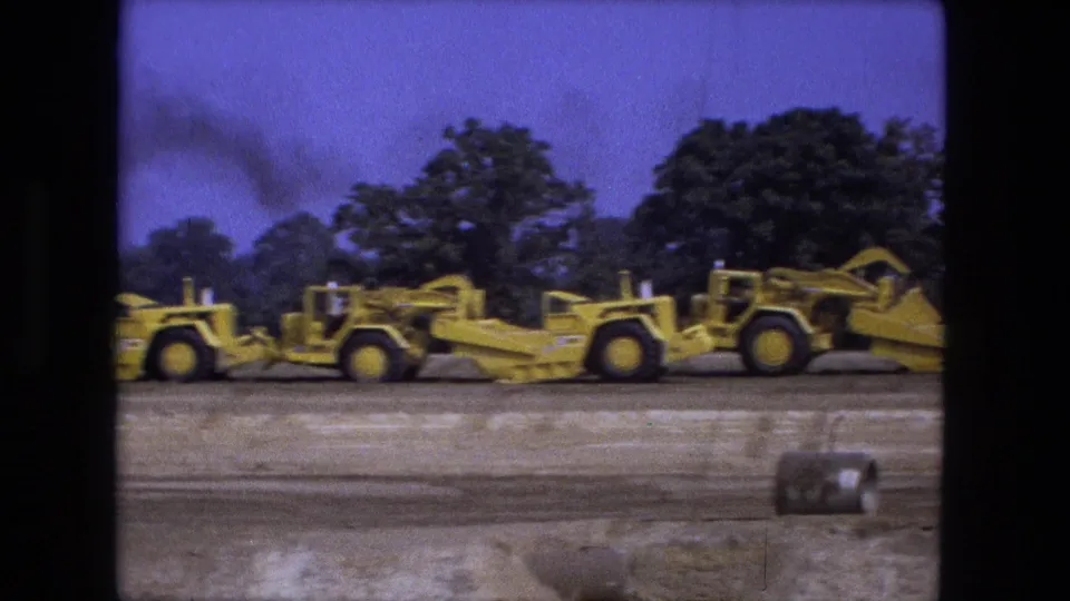 1974 Fort Wayne home-movie frame with yellow grading equipment on a dirt site, trees behind it, and smoke visible against the sky.