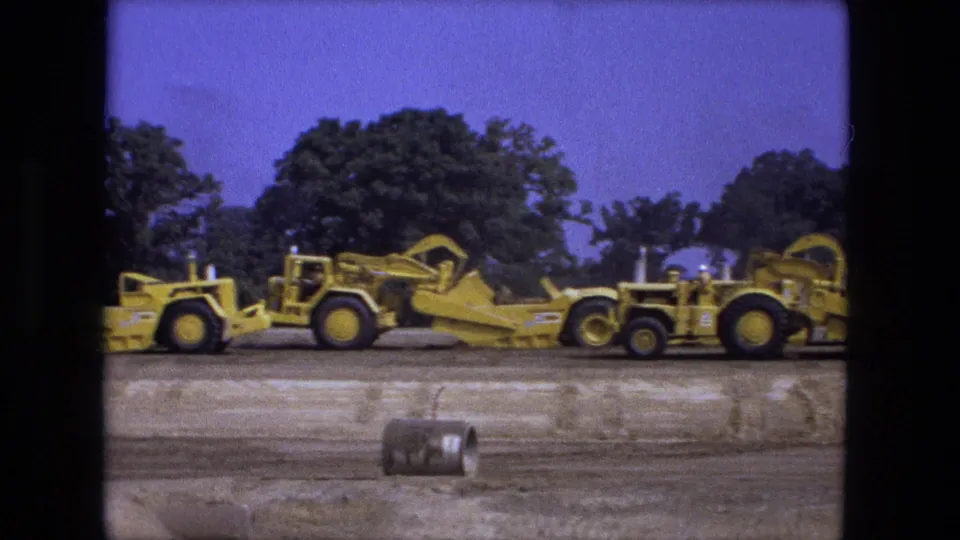 1974 Fort Wayne 8mm frame showing yellow earth-moving machinery, scraped soil, a tree line, and a small barrel-like object in front.
