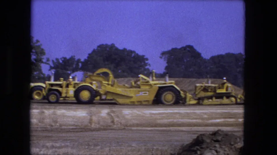 1974 Fort Wayne 8mm frame with a long yellow earth mover, trailing equipment, dirt mounds, trees, and saturated blue sky.