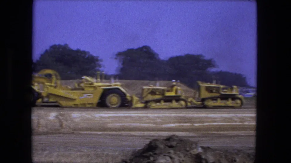 Fort Wayne, Indiana 1974 8mm frame showing yellow ground-leveling machinery crossing a dirt worksite under a blue sky.