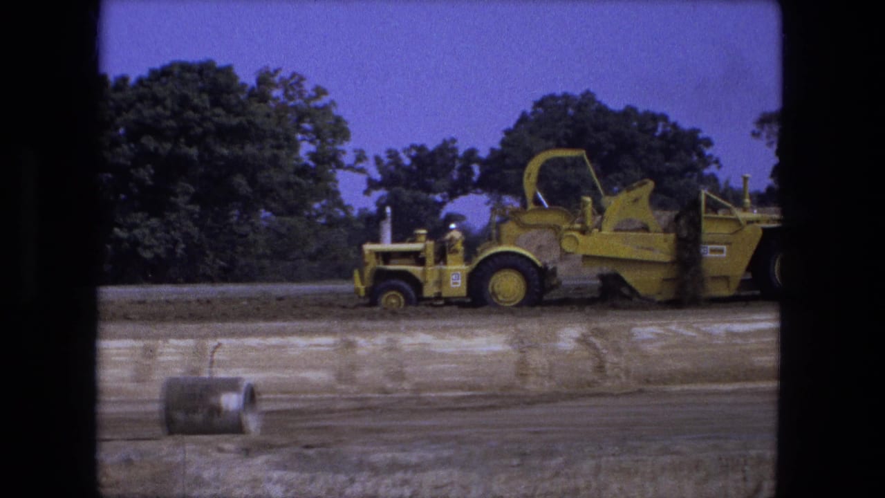 A yellow motor scraper or grader stands on dirt near trees, with a barrel in the foreground.
