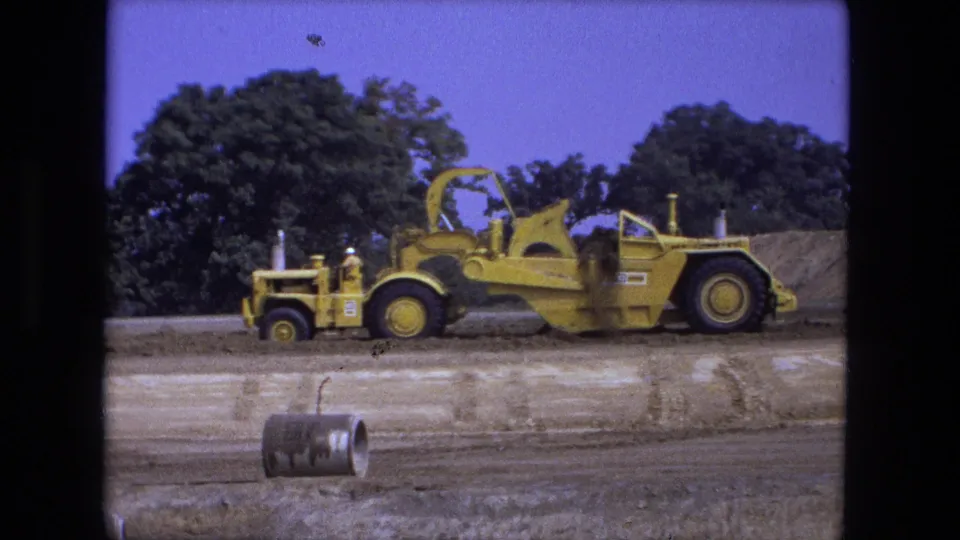 Fort Wayne, Indiana 1974 8mm frame showing a yellow earthmoving machine viewed side-on at a worksite.