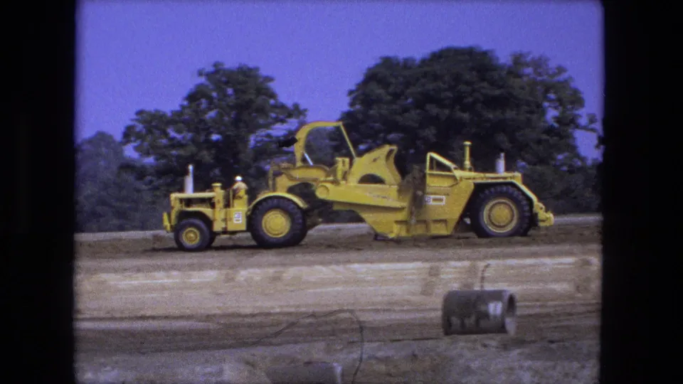 1974 Fort Wayne, Indiana 8mm frame showing a yellow construction machine on graded dirt with trees in the background.