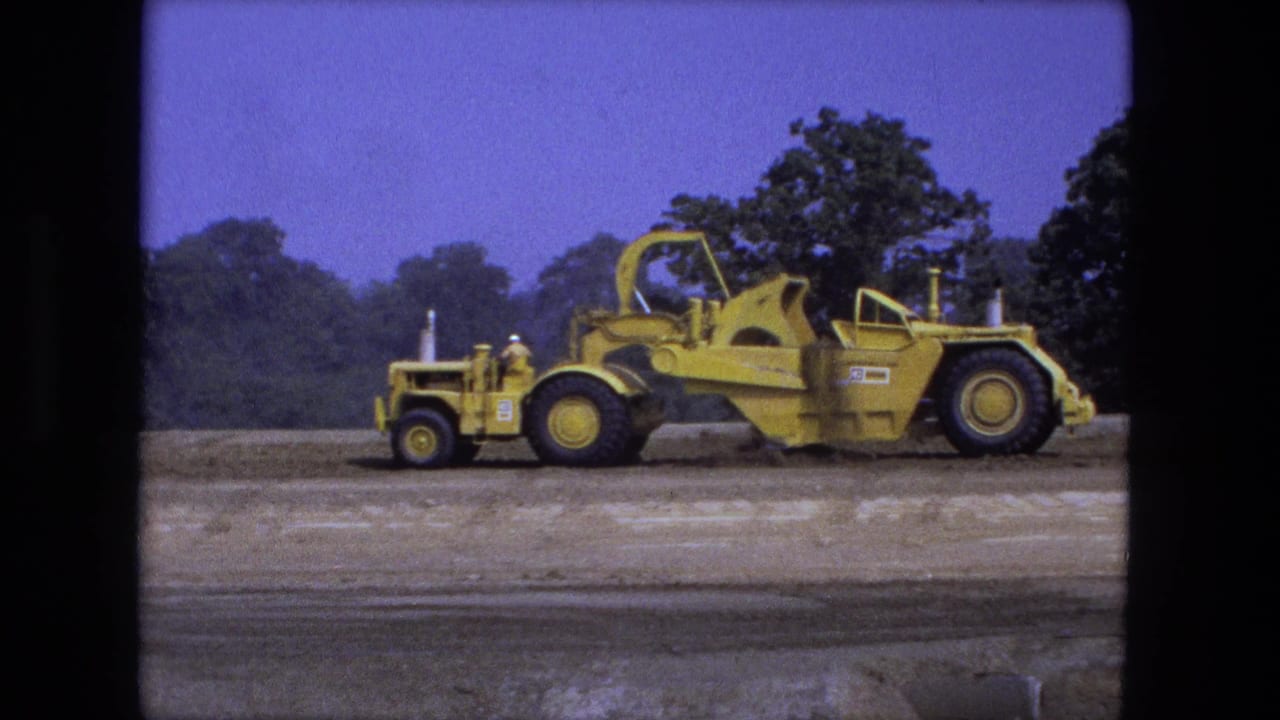 Yellow road grader or scraper crossing a dirt field below a tree line.