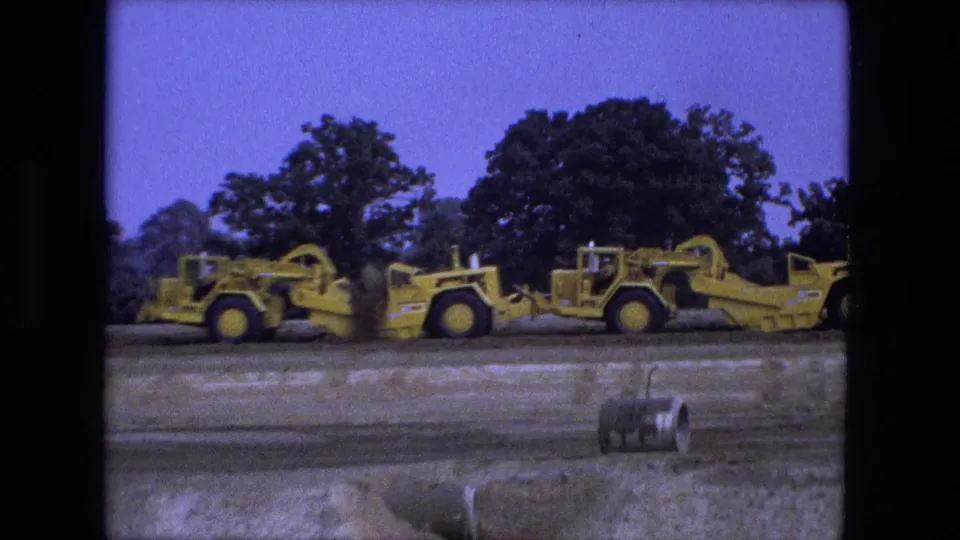 Two yellow earthmoving machines move along a graded rural site near Fort Wayne, Indiana, in 1974 home-movie footage.