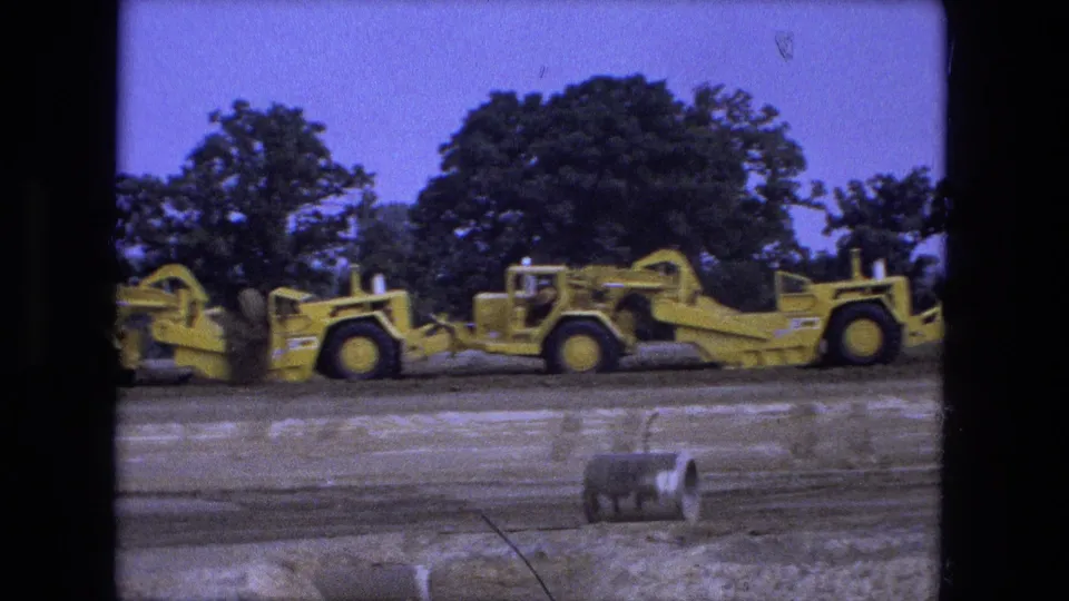 1974 Fort Wayne 8mm frame showing yellow road graders on a dirt work site with operators and a tree line in the background.