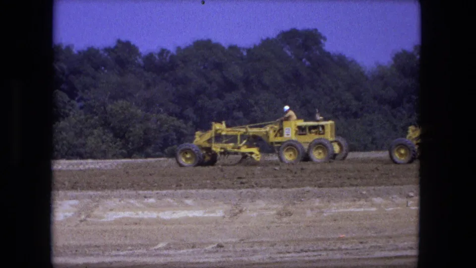 A single yellow road grader crosses a bare construction field in Fort Wayne, Indiana, with another machine partly visible at the edge.