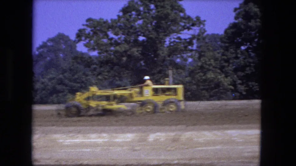 1974 Fort Wayne 8mm frame showing a yellow road grader crossing scraped soil with a tree line in the background.