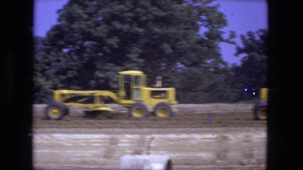 Yellow road grader passes across graded soil beside trees and a partial vehicle at the edge.