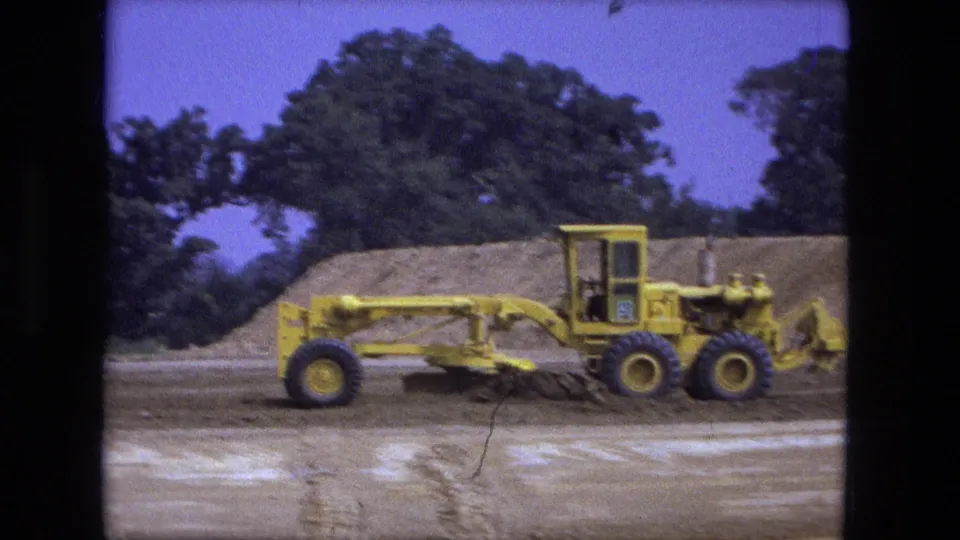 Side view of a yellow road grader at a Fort Wayne construction site, with scraped ground and a dirt mound behind it.