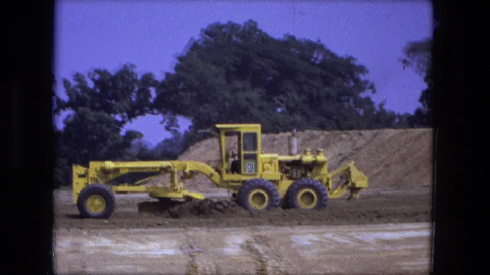 1974 Fort Wayne 8mm frame showing a yellow road grader and operator cab against a dirt mound and trees.