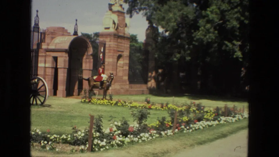 1974 India home-movie frame showing a mounted ceremonial guard beside a formal red stone gateway, lawn, trees, and planted flowers.
