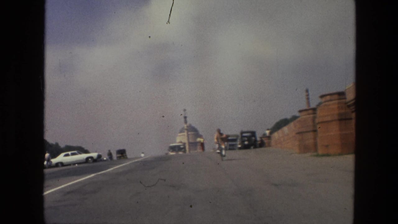 1974 India street view with a broad paved road, classic cars, a cyclist, and domed red sandstone architecture in the distance.