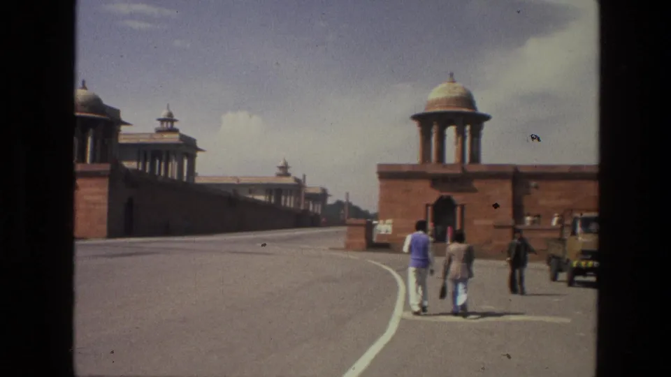 1974 India home-movie frame of pedestrians on a wide roadway bordered by domed red sandstone buildings and walls.