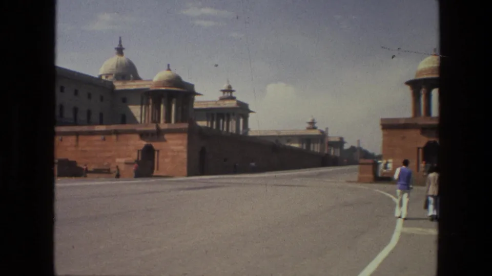 1974 India street scene showing pedestrians at the edge of a broad road with monumental red sandstone buildings under a bright sky.