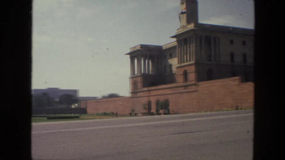 1974 India home-movie frame of a monumental sandstone civic building, landscaped verge, road, and distant pedestrians.
