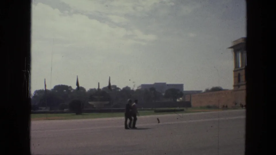 A couple walks through a 1974 India civic-space view, with flagpoles and trees beyond the open roadway.