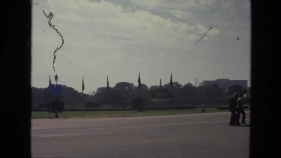 1974 India home-movie frame with a couple at the roadside, flagpoles, lawns, and distant public buildings.