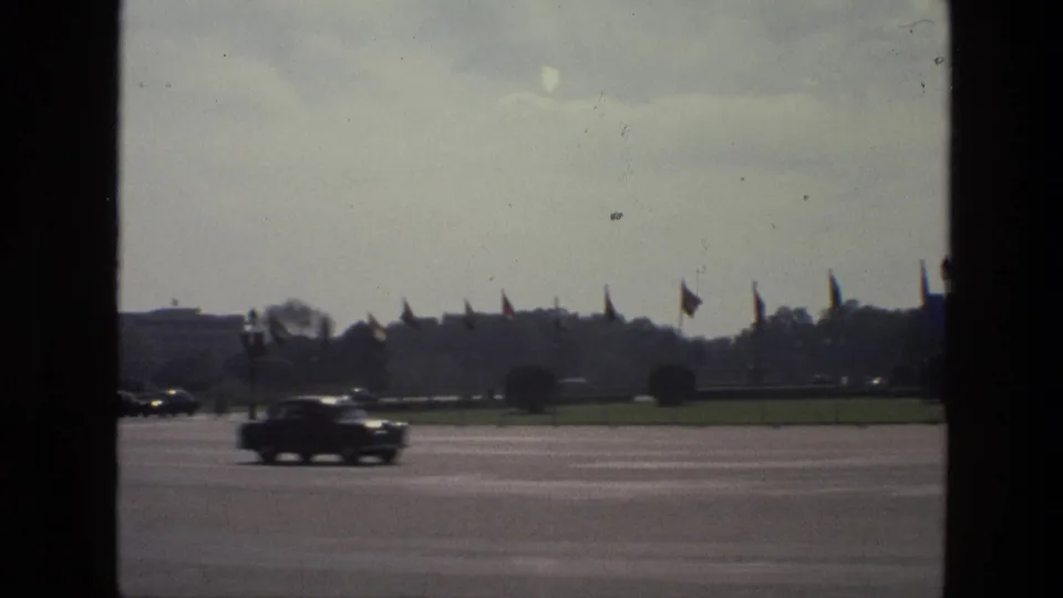 1974 India home-movie frame of a broad city road with passing cars, distant flags, a green median, and dark window framing.