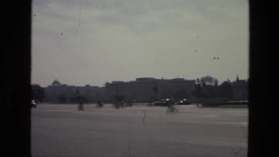 1974 India street scene showing a broad road, distant cyclists and cars, institutional-looking buildings, and window-edge framing.