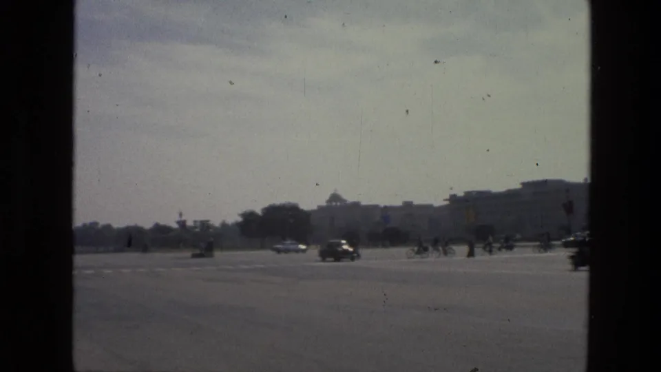 1974 India home-movie frame of cyclists and cars moving along a broad roadway with distant buildings and flags under a pale sky.