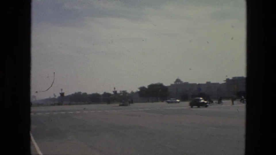 1974 India 8mm frame showing a broad open roadway, passing cars, lawns, and distant institutional buildings in a large public square.