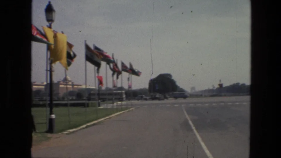 A 1974 India 8mm frame looks along a road bordered by national flags, a lamppost, green lawn, and distant civic buildings.