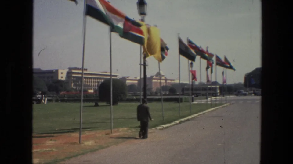 1974 India 8mm frame showing a uniformed man walking past flagpoles, lawns, a broad road, and large buildings in the distance.